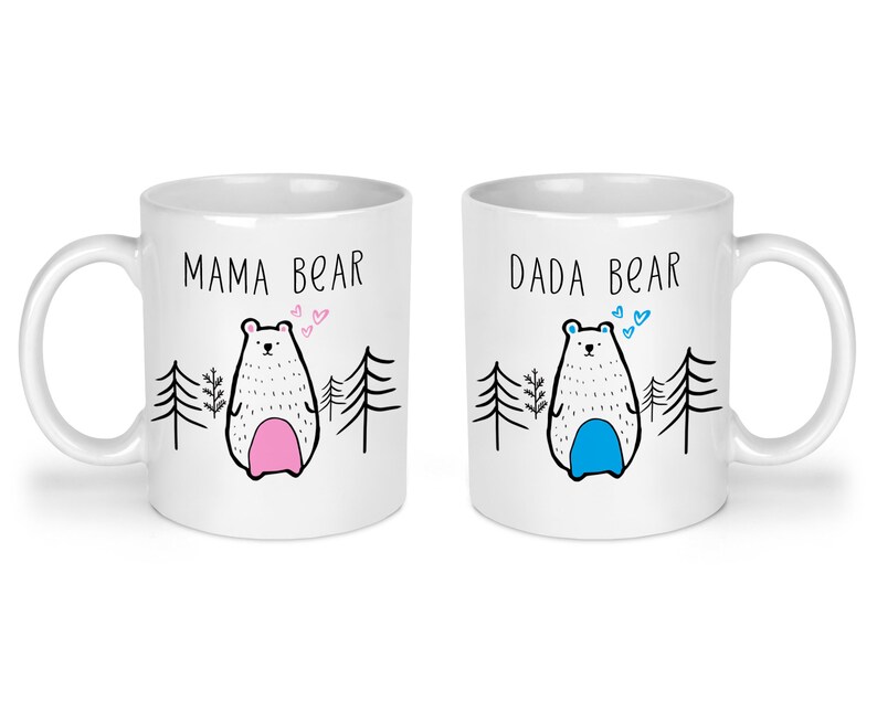 May include: Two white ceramic mugs with black text and illustrations. The mug on the left says "Mama Bear" and features a white bear with pink accents. The mug on the right says "Dada Bear" and features a white bear with blue accents.