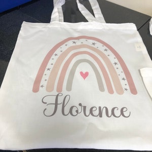K&ouml;nnte beinhalten: Wei&szlig;e Stofftasche mit einem Regenbogen-Design in Rosa, Beige und Grau. Der Regenbogen ist mit kleinen grauen Sternen und einem pinkfarbenen Herzen in der Mitte verziert. Der Name "Florence" ist in Grau unter dem Regenbogen aufgedruckt.