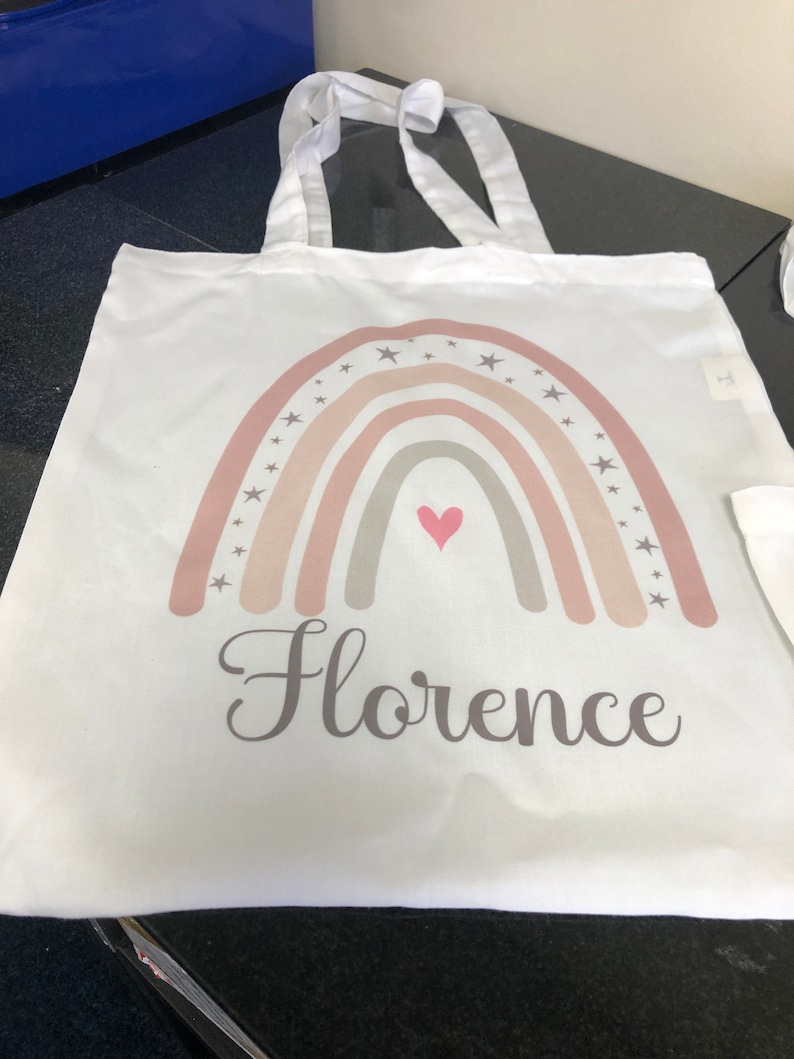 K&ouml;nnte beinhalten: Wei&szlig;e Stofftasche mit einem Regenbogen-Design in Rosa, Beige und Grau. Der Regenbogen ist mit kleinen grauen Sternen und einem pinkfarbenen Herzen in der Mitte verziert. Der Name "Florence" ist in Grau unter dem Regenbogen aufgedruckt.