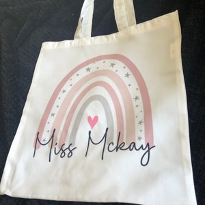 K&ouml;nnte beinhalten: Wei&szlig;e Stofftasche mit einem rosafarbenen, grauen und wei&szlig;en Regenbogenmuster und einem rosafarbenen Herzen. Der Text "Miss McKay" ist unter dem Regenbogen gedruckt.