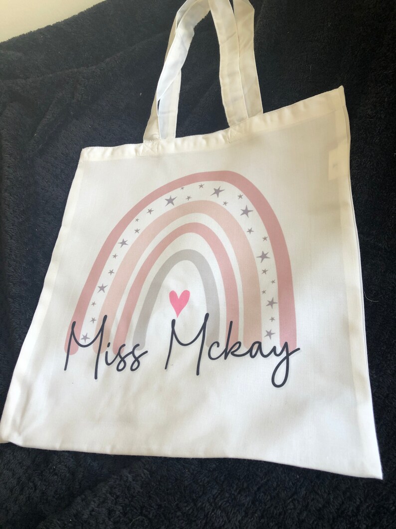 K&ouml;nnte beinhalten: Wei&szlig;e Stofftasche mit einem rosafarbenen, grauen und wei&szlig;en Regenbogenmuster und einem rosafarbenen Herzen. Der Text "Miss McKay" ist unter dem Regenbogen gedruckt.