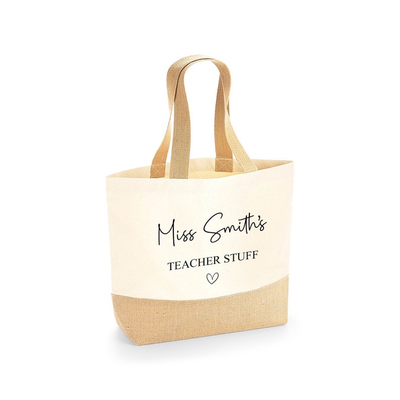 K&ouml;nnte beinhalten: Beige Canvas-Tragetasche mit Juteboden und passenden Griffen. Auf der Tasche steht in schwarzer Schrift "Miss Smith's TEACHER STUFF" mit einem kleinen Herzsymbol darunter, ideal f&uuml;r Lehrer.