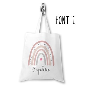 K&ouml;nnte beinhalten: Wei&szlig;e Einkaufstasche mit einem Regenbogen-Design in Rosa, Beige und Grau mit wei&szlig;en Sternen und einem rosa Herzen. Der Name "Sophia" ist unter dem Regenbogen gedruckt.