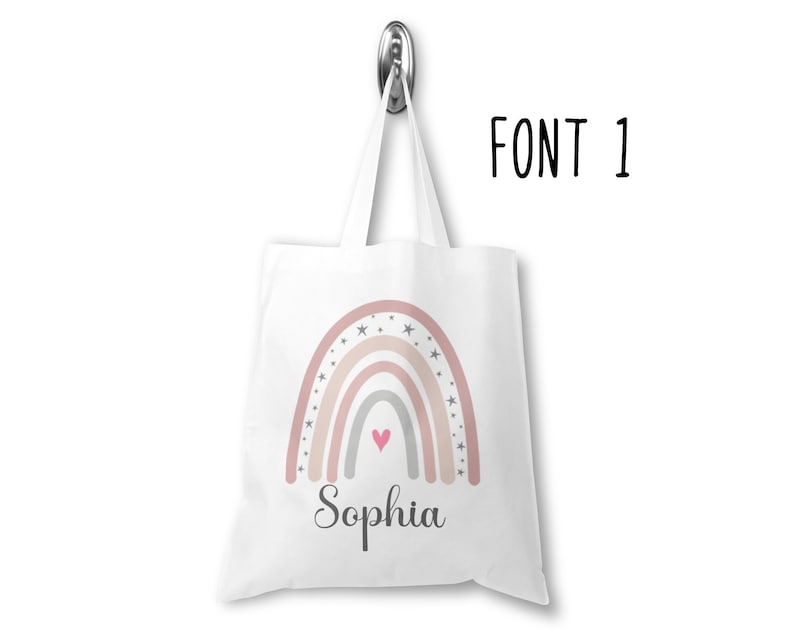 K&ouml;nnte beinhalten: Wei&szlig;e Einkaufstasche mit einem Regenbogen-Design in Rosa, Beige und Grau mit wei&szlig;en Sternen und einem rosa Herzen. Der Name "Sophia" ist unter dem Regenbogen gedruckt.