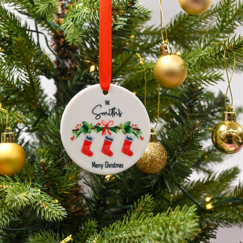 May include: White ceramic Christmas ornament with a red ribbon. The ornament features a green holly wreath with red berries and three red stockings with the names "Daddy", "Mommy", and "Sophia" written on them. The text "The Smith's" and "Merry Christmas" is also printed on the ornament.