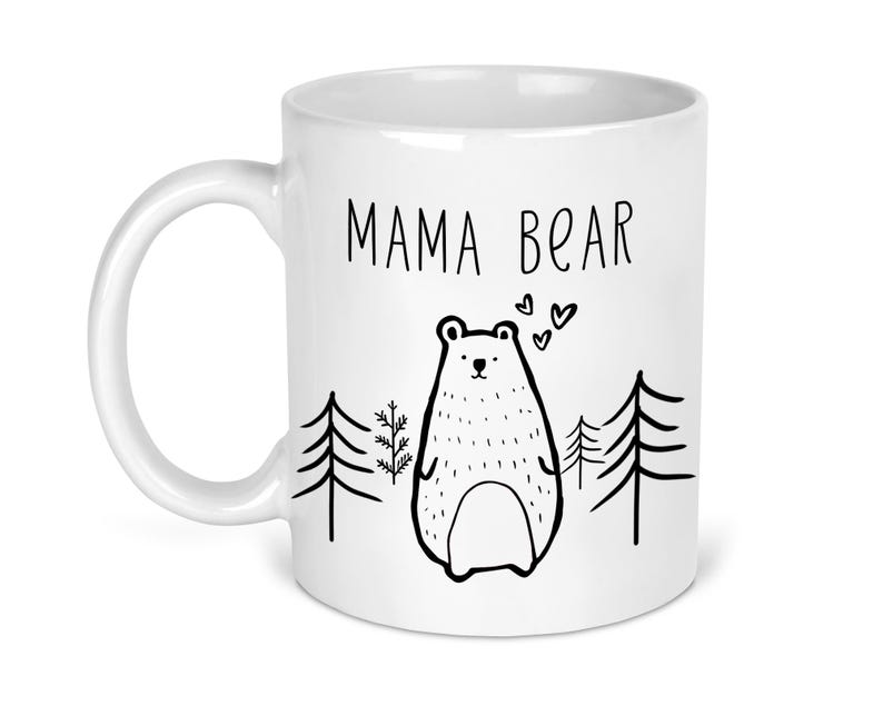 May include: White ceramic mug with a black outline of a mama bear and two pine trees. The text "Mama Bear" is printed in black on the mug.