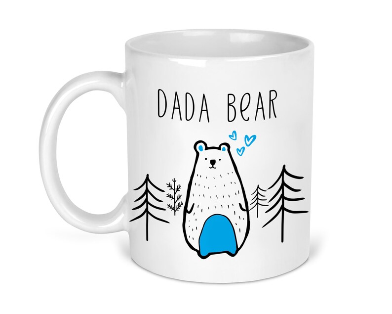 May include: White ceramic mug with a black outline of a bear with blue accents and the text "DADA BEAR" in black. The bear is surrounded by black outlines of pine trees.