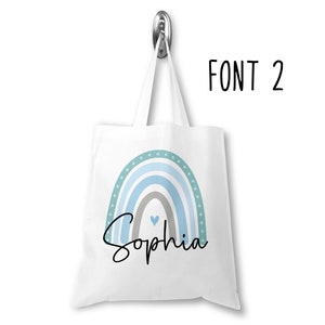 K&ouml;nnte beinhalten: Wei&szlig;e Einkaufstasche mit einem blauen und gr&uuml;nen Regenbogen-Design und dem Namen "Sophia" in schwarzer kursivschrift. Die Tasche hat ein kleines Herz in der Mitte des Regenbogens.