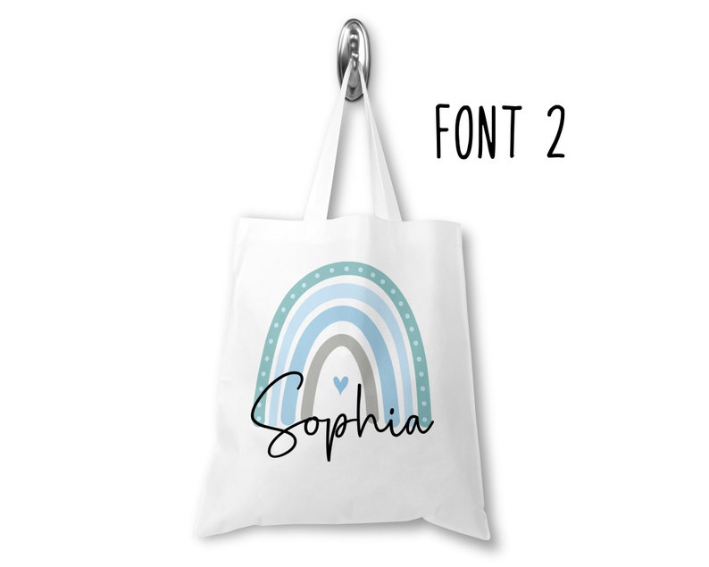 K&ouml;nnte beinhalten: Wei&szlig;e Einkaufstasche mit einem blauen und gr&uuml;nen Regenbogen-Design und dem Namen "Sophia" in schwarzer kursivschrift. Die Tasche hat ein kleines Herz in der Mitte des Regenbogens.