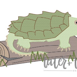 Pode incluir: Design bordado de uma tartaruga-aligátor verde-claro com a boca aberta, pousada num tronco castanho. A tartaruga tem uma carapaça texturizada e é rodeada por erva verde. O texto "tater + tots designs" está por baixo.