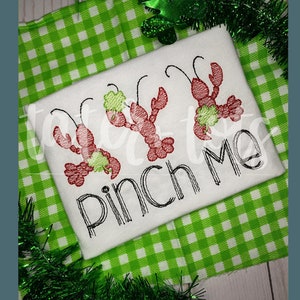 Op de afbeelding: Wit T-shirt met geborduurde rode rivierkreeften met groene klavers. De tekst "Pinch Me" staat in zwart schrift onder de rivierkreeften. Het shirt ligt op een groen-witte geruite achtergrond.