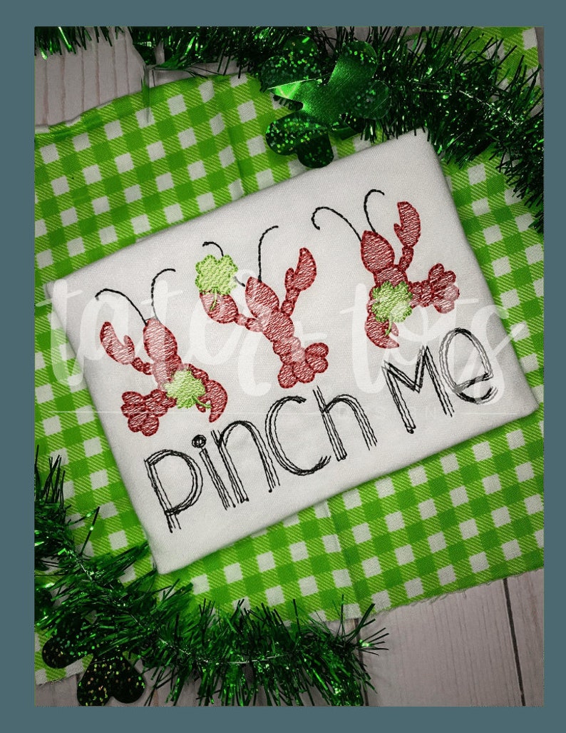 Op de afbeelding: Wit T-shirt met geborduurde rode rivierkreeften met groene klavers. De tekst "Pinch Me" staat in zwart schrift onder de rivierkreeften. Het shirt ligt op een groen-witte geruite achtergrond.