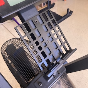 May include: Black metal phone holder with a grid pattern and screws. The holder is attached to a black exercise machine.
