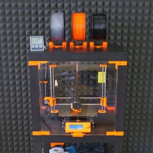 May include: A black 3D printer with an orange frame and a clear plastic enclosure. The printer has a spool holder with three spools of filament: gray, orange, and black. A digital thermometer is mounted on the top of the printer.