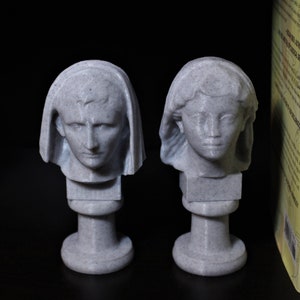 May include: Two gray stone busts of people with draped fabric on their heads. The busts are on small gray stone bases. The busts are in front of two books with yellow covers. The book on the left has the title "The Birth of Politics" and the author "Lane" printed on the cover. The book on the right has the title "A History of Ancient Rome" printed on the cover.