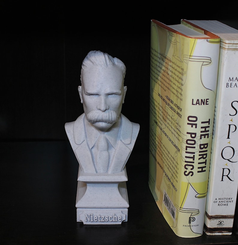 Puede incluir: Un busto de yeso blanco de Friedrich Nietzsche, con bigote, vestido con traje y corbata. El busto est&aacute; sobre una peque&ntilde;a base con la palabra "Nietzsche" inscrita en ella. El busto est&aacute; junto a dos libros, uno titulado "El nacimiento de la pol&iacute;tica" y el otro titulado "SPQR: Una historia de la antigua Roma".