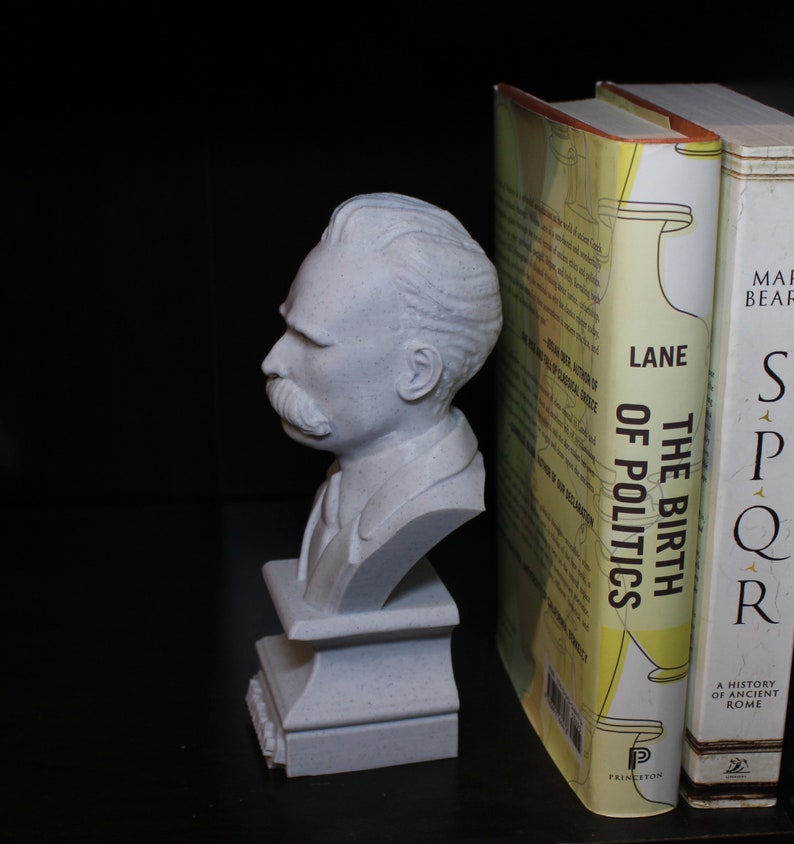 Puede incluir: Un busto de yeso blanco de un hombre con bigote, sobre un pedestal blanco. El busto se utiliza como sujetalibros para dos libros. El libro de la izquierda se titula "The Birth of Politics" de Lane.