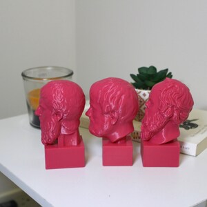 Puede incluir: Tres bustos de hombres con barba impresos en 3D de color rosa. Los bustos est&aacute;n dispuestos en fila sobre una superficie blanca.