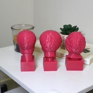 Puede incluir: Tres bustos de personas con pelo corto, impresos en 3D en color rosa, que se encuentran sobre peque&ntilde;as bases cuadradas. Los bustos est&aacute;n dispuestos en fila sobre una superficie blanca.