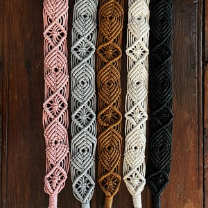 May include: Five macrame straps in different colors, including pink, gray, brown, white, and black. Each strap has a gold metal clasp.
