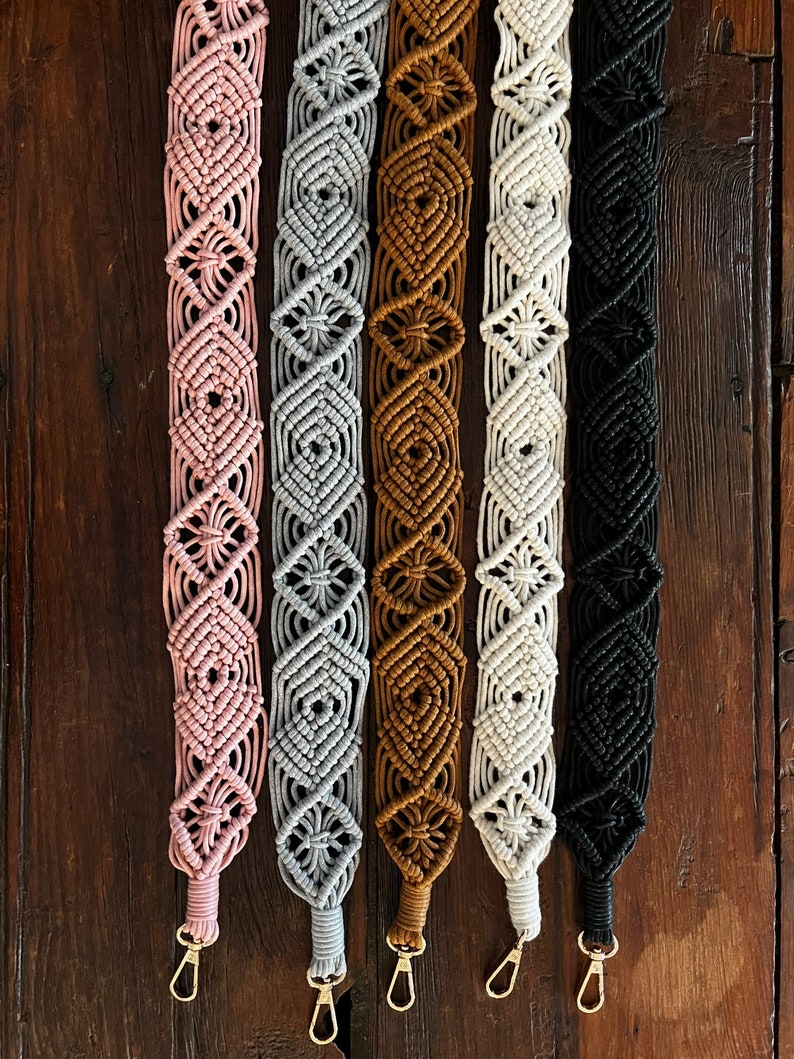 May include: Five macrame straps in different colors, including pink, gray, brown, white, and black. Each strap has a gold metal clasp.