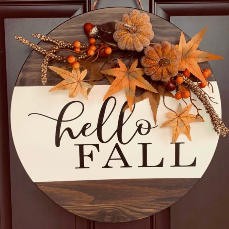 May include: A round wooden door hanger with a white and brown wood grain pattern. The top half of the door hanger is decorated with fall-themed foliage, including orange and brown leaves, small orange berries, and two brown and gold glitter pumpkins. The bottom half of the door hanger is white with the words 'hello fall' written in black cursive font.