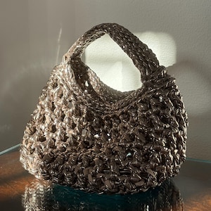 May include: A brown metallic woven handbag with a rounded shape and a single handle. The bag features an open weave design, and the handle is also woven. The bag is placed on a reflective surface.