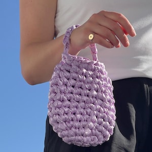 Puede incluir: Un bolso de mano de color lavanda tejido a mano con un aspecto texturizado y tejido. El bolso tiene una forma redondeada con un asa a juego. El fondo es un cielo azul claro, lo que sugiere un entorno al aire libre. El bolso lo sostiene una persona.