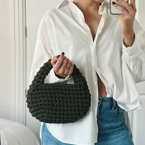Puede incluir: Un bolso de mano de crochet de color verde oscuro con forma redondeada. El bolso lo lleva una persona que lleva una camisa blanca abotonada y pantalones vaqueros azules.