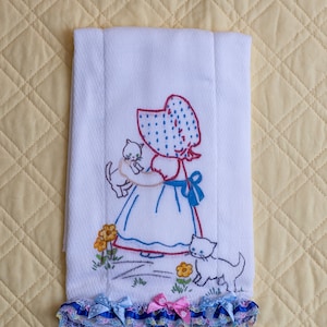 Puede incluir: Toalla de cocina blanca bordada con una niña con un sombrero rojo con lunares blancos que sostiene un gatito blanco. La niña lleva un vestido azul y un lazo azul. Hay dos gatitos blancos en la toalla, uno en los brazos de la niña y otro en el suelo. La toalla tiene un ribete de volantes azules, blancos y rosas.
