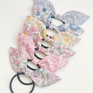 May include: A set of five floral print hair scrunchies with black elastic bands. The scrunchies are made of cotton fabric and feature a variety of floral patterns in shades of pink, blue, yellow, and green.