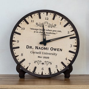 Puede incluir: Un reloj redondo de madera con marco y soporte marrón oscuro. La esfera del reloj es de color claro con números y manecillas negras. El texto en la esfera del reloj dice "DR. NAOMI OWEN Cornell University MAY 2020" y una cita. Decoración del hogar.