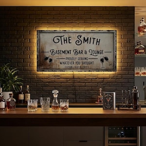 May include: A rectangular wall sign with a distressed finish, featuring the text THE SMITH BASEMENT BAR & LOUNGE, PROUDLY SERVING WHATEVER YOU BROUGHT, and ESTABLISHED 2023. The sign is illuminated by a warm LED backlight against a dark brick wall.