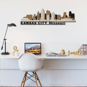 May include: A wooden wall art piece depicting the Kansas City skyline with the text "KANSAS CITY Missouri" below. The art is displayed on a white wall above a white desk with a laptop, a white chair, and a gold "smile" sign.