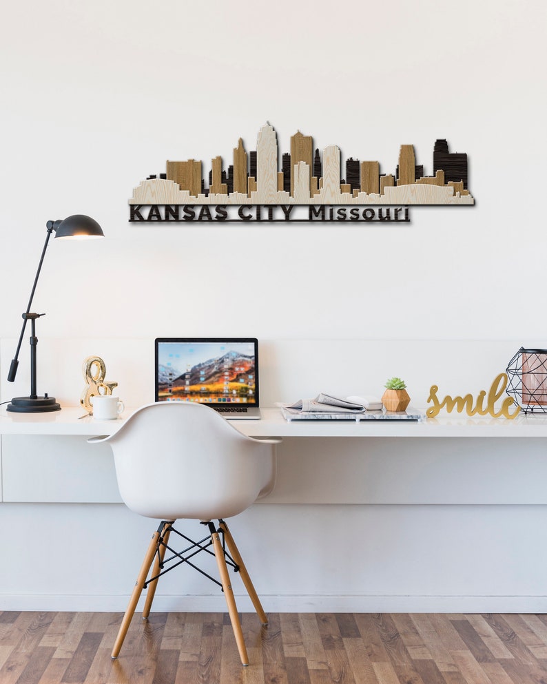 May include: A wooden wall art piece depicting the Kansas City skyline with the text "KANSAS CITY Missouri" below. The art is displayed on a white wall above a white desk with a laptop, a white chair, and a gold "smile" sign.