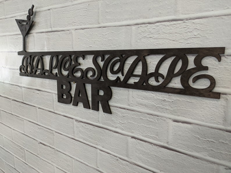 May include: A black wooden sign with the words "BLUESCAPE BAR" in a decorative font. A small martini glass with olives is above the words.