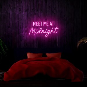 May include: Pink neon sign with the text "Meet me at Midnight" hanging on a dark wood wall above a red bed with pillows.