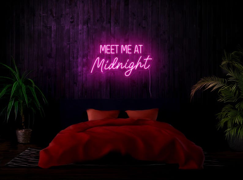 May include: Pink neon sign with the text "Meet me at Midnight" hanging on a dark wood wall above a red bed with pillows.