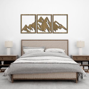 Pu&ograve; includere: Tre pezzi di arte da parete in legno che raffigurano una catena montuosa in stile minimalista. I pezzi sono disposti orizzontalmente e sono montati su una parete bianca sopra un letto con un copriletto grigio.