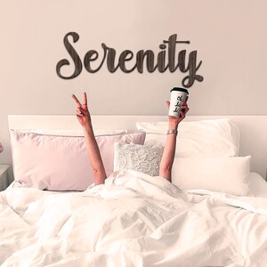 May include: A white bed with white bedding and pink pillows. The word "Serenity" is written on the wall above the bed in brown letters. A person is lying in the bed with their arms raised, holding a white coffee cup with the words "oh la la" written on it.