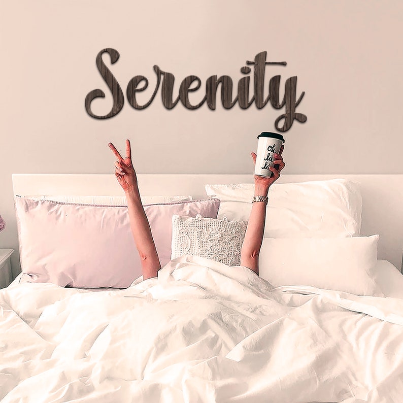 May include: A white bed with white bedding and pink pillows. The word "Serenity" is written on the wall above the bed in brown letters. A person is lying in the bed with their arms raised, holding a white coffee cup with the words "oh la la" written on it.