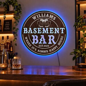 Könnte beinhalten: Ein rundes Wand-Schild aus Holz mit blauer LED-Hintergrundbeleuchtung, das den Text "WILLIAM'S BASEMENT BAR" und "WHERE IT'S ALWAYS HAPPY HOURS", eine Abbildung von zwei anstoßenden Bierkrügen sowie "EST. 2025" in weißer Schrift zeigt.