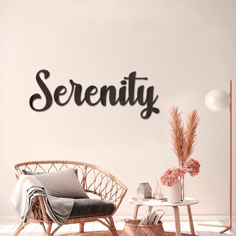 May include: A wooden wall decor sign with the word "Serenity" in a cursive font. The sign is brown and is hanging on a white wall. The sign is in a living room setting with a wicker chair, a side table, and a floor lamp.