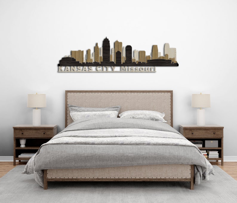 May include: A wood wall art piece depicting the Kansas City skyline with the text "KANSAS CITY Missouri" below. The art is displayed above a bed with a grey duvet cover and two matching bedside tables with lamps.