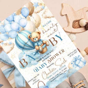 Puede incluir: Una invitación de baby shower azul y dorada con un osito de peluche en un globo aerostático. La invitación dice "We can bearly wait!" y "Baby Shower" con el nombre "Charlotte Smith".