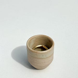 Puede incluir: Una pequeña taza cilíndrica de cerámica con un acabado mate en color beige claro y sutiles estrías horizontales en la parte inferior. Dentro de la taza descansa un anillo de metal dorado fino con una superficie texturizada y granulada.