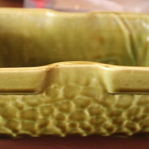 以下が含まれることがあります： 表面がざらざらとした質感の緑色の陶器製のプランター。プランターは角が丸みを帯びた正方形の形をしています。