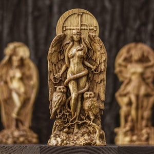 May include: A wooden carving of a winged female figure with a snake wrapped around her. She is standing on a base with two owls on either side. The carving is painted in shades of brown and gold.