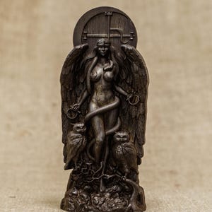 Puede incluir: Una talla de madera oscura de una figura femenina alada con una serpiente enroscada a su alrededor. Ella est&aacute; de pie sobre una base con dos b&uacute;hos a cada lado. La talla es detallada e intrincada.