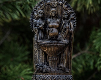 May include: A wooden carving of the Triple Goddess, a representation of the three phases of the feminine divine. The carving features three figures, each with a different expression and pose, standing around a cauldron. The carving is dark brown and has a detailed, intricate design.